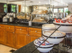 Breakfast buffet with cereals, coffee, bagels, breads, and condiments on a counter. Wooden cabinetry below and toasters visible in the background.