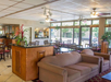 A cozy seating area with a sofa and chairs is in the foreground. Tables and chairs fill the dining area. Large windows let in natural light, and plants add a touch of greenery.