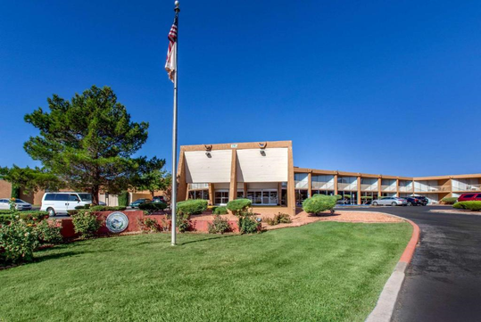 Building with triangular entrance, surrounded by landscaping and cars. An American flag is on a pole in front. The sky is clear and blue.