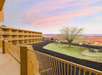View from a balcony overlooking a hotel courtyard with a tree, picnic tables, and distant hills under a cloudy sky at sunset.