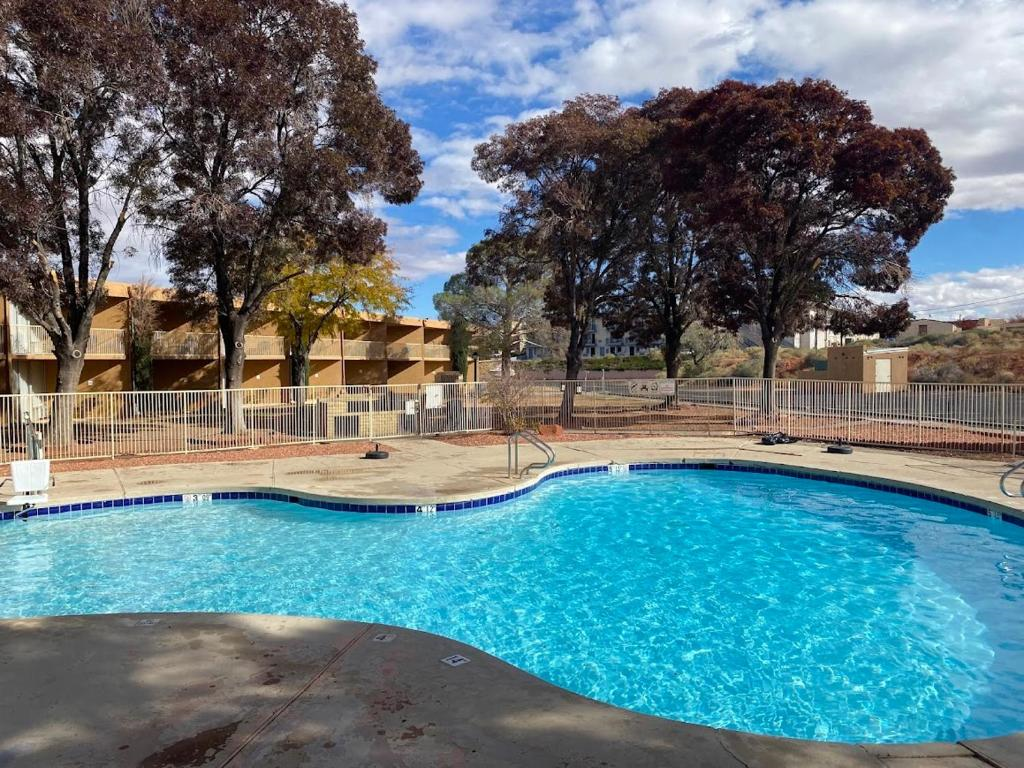 Outdoor swimming pool with clear water, surrounded by a concrete deck and white metal fence. Trees and buildings visible in the background under a partly cloudy sky.