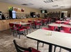 A cafeteria-style dining area with empty tables and red chairs, beverage dispensers, and a counter along the back wall.