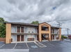 Two-story motel building with exterior corridors, stairway in the center, several doors and windows, and an empty parking lot in front under a cloudy sky.