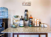 Coffee station with hot water, regular and decaf coffee dispensers, a water cooler, bowls, cups, and lids on a granite countertop.