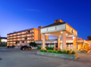 Hotel exterior at dusk with a well-lit entrance, multiple floors, a parking area with a car, and landscaped greenery.