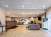 Hotel lobby with seating area, check-in desk, banner display, and American flag. Decor includes circular wall art. Bright lighting with tiled floor and ceiling grid.