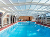 Indoor swimming pool with a transparent roof, surrounded by red-tiled flooring. Lounge chairs and tables are placed along the sides.