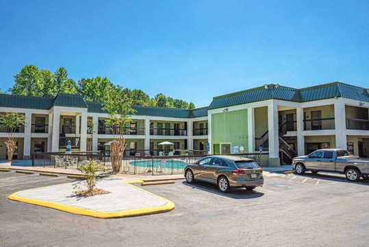 Two-story motel with a green roof and a central outdoor pool. Cars are parked in the front, and trees are in the background.