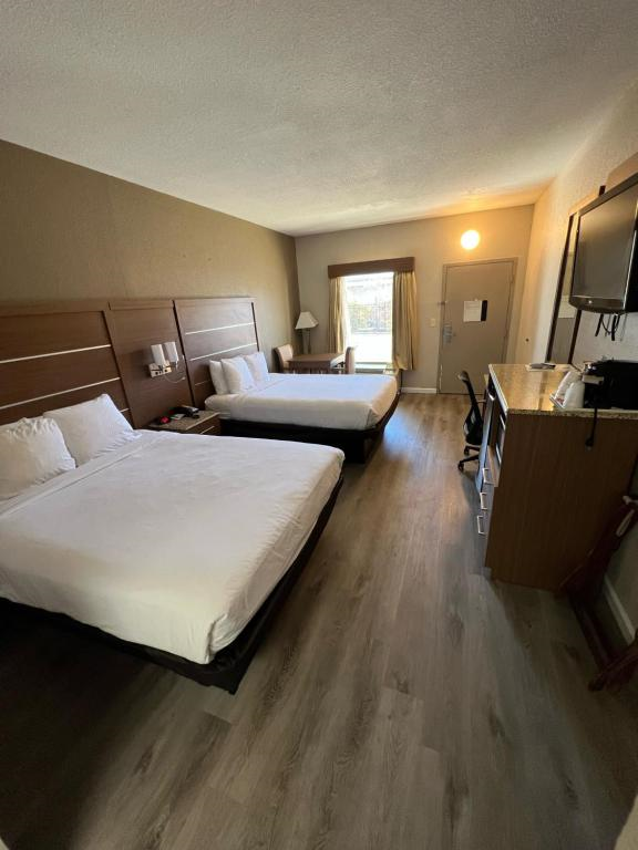 Hotel room with two double beds, a flat-screen TV, desk, and chair. Hardwood floor, brown and white decor. Window with curtain. Door near the beds.