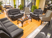 Spacious living room with black leather sofas, patterned armchairs, and a wooden coffee table on a wooden floor. Large windows and indoor plants are in the background.