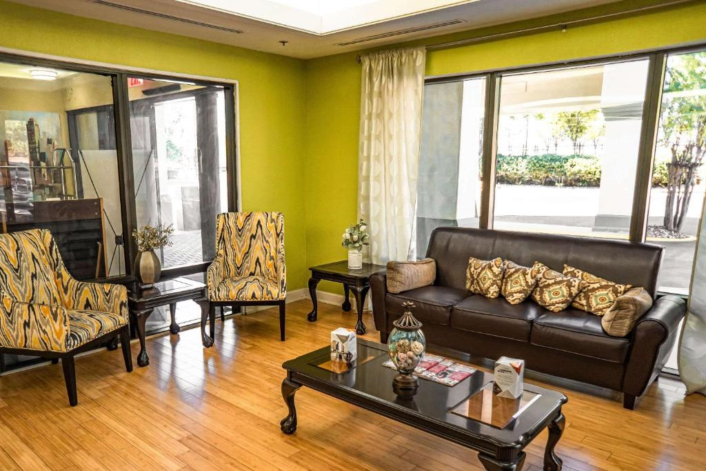 A modern hotel lobby with green walls, large windows, a leather sofa with patterned cushions, two patterned chairs, and dark wood coffee tables on a wooden floor.