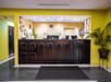 Hotel reception area with a dark wooden front desk, plants, and a sign reading "Quality Inn & Suites" on the wall. Bright yellow walls and tiled floor with a black mat in front of the desk.