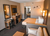 A hotel room with a double bed, sofa, desk, and TV. Includes a mini-fridge, two mirrors, wall art, and a bathroom visible in the background. The decor is neutral with brown tones.