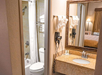 Bathroom with a sink, mirror, and wall-mounted hairdryer in the foreground. A small bathtub with a shower curtain and toilet are visible through an open door in the background.