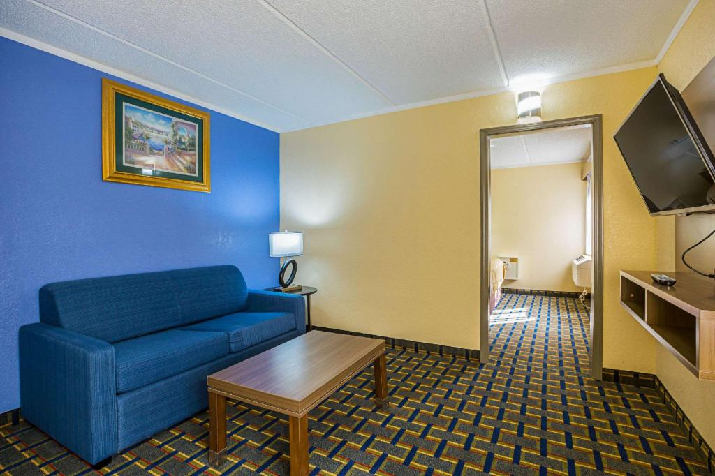Hotel room with a blue couch, wooden coffee table, wall-mounted TV, and a lamp. The walls are blue and yellow, with a framed painting. A doorway leads to another room.