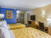 Hotel room with two double beds, a TV on a wooden desk, a chair, and wall-mounted lamps. A framed painting is on one wall, and a mirror is on the other. Carpeted floor and beige-blue walls.