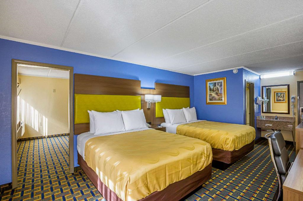 Hotel room with two double beds, yellow bedspreads, blue walls, wall art, a mirror, and a desk with a chair. Bright lighting and carpeted floor.
