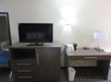 Hotel room with a TV on a wooden dresser, a desk with a lamp, a phone, and a coffee maker. A mirror is on the left wall, and a wastebasket is beneath the desk.
