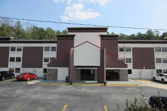 A three-story building with a modern facade and flat roof, featuring multiple windows and a central entrance. Several cars are parked in the front lot. Trees are visible behind the building.