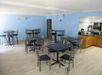 A room with light blue walls featuring multiple black tables and gray chairs. A countertop with a coffee machine and cup dispenser is visible on the right.