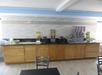 Breakfast buffet with cereal dispensers, juice machine, and coffee makers on a counter. Wooden cabinets below and a textured white wall behind.