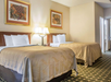 Hotel room with two double beds, gold bedspreads, white pillows, two wall paintings above the beds, and a lamp in between. Carpeted floor and open door leading to another room.