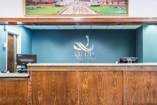 Front desk of Quality Inn & Suites with two computer monitors and a wooden counter, under a painting on the blue wall.