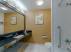 A modern bathroom with a long mirror, sink, toilet, and shower. Walls are yellow with white tiles on the floor. A framed abstract artwork hangs above the toilet.