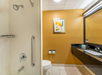 Bathroom with a walk-in shower, safety grab bar, toilet, wall artwork, and a long countertop with a sink and toiletries. Walls are painted yellow.