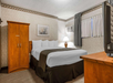 A hotel room with a double bed, white bedding, and a dark throw. Features include a wooden wardrobe, a lamp, an air conditioner, and a TV on a wooden dresser. Artwork hangs on the wall.