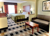 A hotel room with a bed, a sofa, a wooden coffee table, a desk with a chair, two lamps, and a window with curtains. The room has patterned carpeting and a framed artwork on the wall.