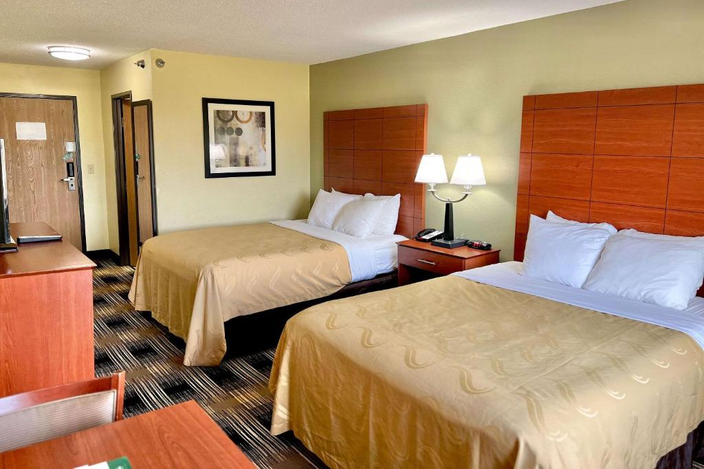 Hotel room with two double beds, wooden furniture, a table lamp, and wall art. The carpet is patterned and the walls are painted yellow. A door is visible in the background.