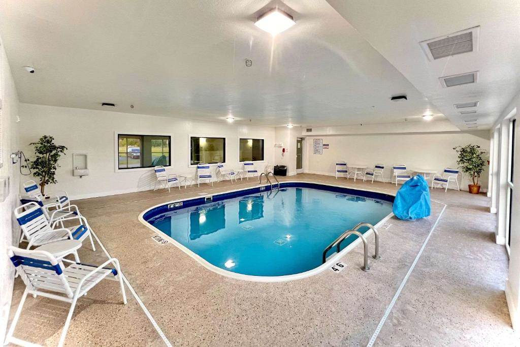 Indoor swimming pool with blue water, surrounded by white lounge chairs and green plants. Bright overhead lighting, windows, and safety notices on the walls.