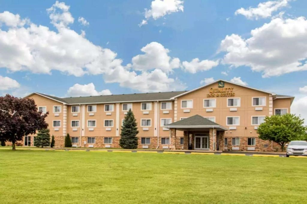 Exterior view of a three-story hotel with a tan facade, surrounded by green lawn and trees, under a partly cloudy sky.