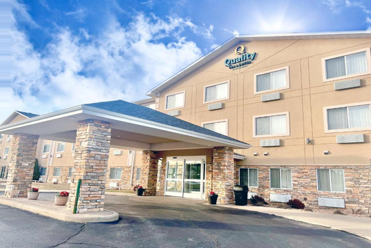 A three-story hotel building with a covered entrance, stone accents, and the "Quality Inn" logo on the facade. Bright blue sky with some clouds in the background.