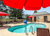 A small outdoor swimming pool with clear water and several lounge chairs and table with red umbrellas around it on a bright sunny day.