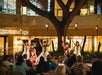 A group of hula dancers in traditional attire performs with paddles in an outdoor setting, watched by an audience.