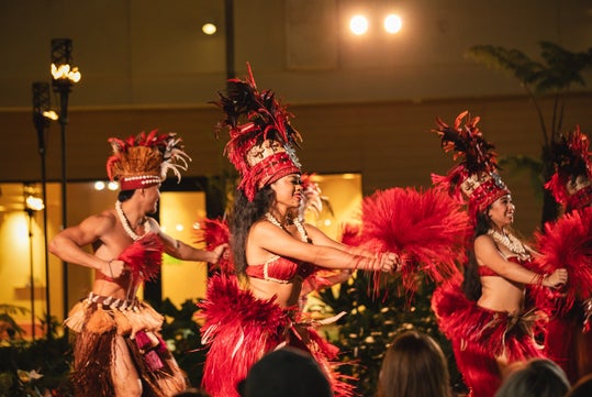 Dancers perform in traditional Polynesian attire, featuring red and brown feathered costumes, on a stage with decorative lighting.