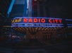 Marquee of Radio City Music Hall lit up at night, with bright red and blue neon lights against a backdrop of tall buildings in an urban setting.