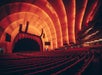 A large, empty concert hall with red seats and warm lighting. The stage is covered by a curtain, and the ceiling features curved, illuminated panels.