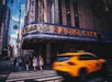 Yellow taxi passes by Radio City Music Hall marquee in New York City. Pedestrians cross the street under a blue sky.