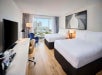 Hotel room with two double beds, a desk with a lamp, a TV, and a city view window. There’s artwork above the beds and a blue chair near the window.