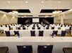 Conference room with rows of tables and chairs facing a projector screen. Tables are set with water bottles and glasses. Ceiling lights illuminate the space.