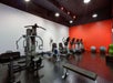 A modern gym with various exercise equipment, including treadmills, stationary bikes, a rowing machine, and weightlifting machines. A bright red accent wall and fitness balls are visible.