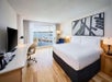 Modern hotel room with a large bed, desk, and chair. A window offers a view of a river and boats. A framed abstract artwork hangs above the bed.