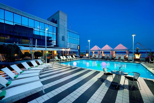 A rooftop pool area with lounge chairs, a striped tile deck, and illuminated spherical lights. The building has reflective glass exteriors. Tents are visible in the background.