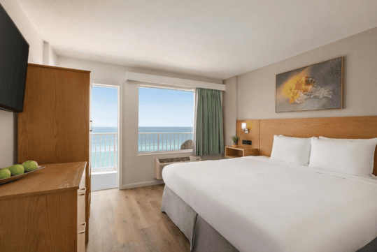 Hotel room with a large bed, wooden furniture, wall-mounted TV, beach view from the window, and a painting above the bed.