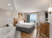 Hotel room with a large bed, a wall-mounted TV, a desk with chairs, a whirlpool tub, and a window showing an ocean view.