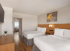 Hotel room with two double beds, wooden flooring, a wall-mounted TV, and a painting above the beds. A small potted plant sits on a dresser, and there's an open doorway to a seating area.
