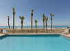 Swimming pool overlooking a beach with rows of palm trees and blue umbrellas in the background.